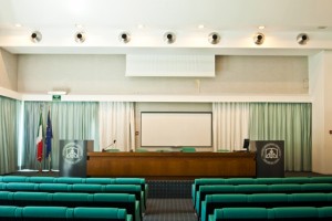 l'aula magna del collegio, vista dalla platea: le poltrone verdi e il tavolo di legno dei relatori; ai lati due podi grigi con il logo del Collegio